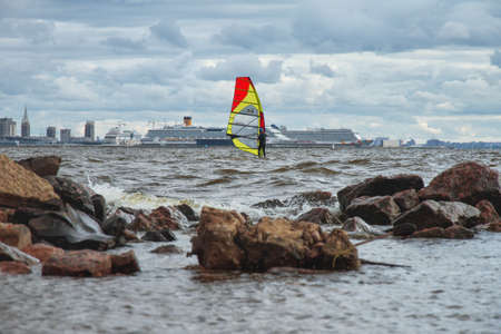 St. Petersburg, Russia, August 24, 2019: male sportsman with red yellow sail on sailing board goes in for sports windsurfing on waves of Gulf of Finland opposite the cruise liners in the portのeditorial素材