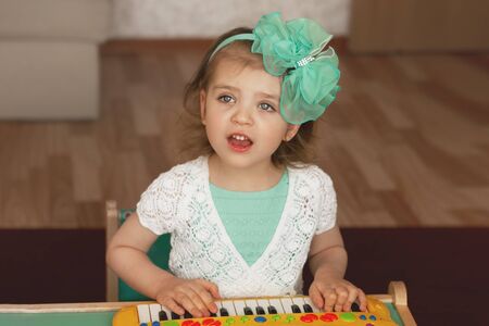 portrait of cheerful girl, plays childrens piano and sings in green dress with white lace and bow on her head selective focusの写真素材
