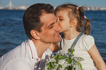 little child girl daughter kisses her young dad on background of sea on sunny day and gives bouquet of flowersの写真素材