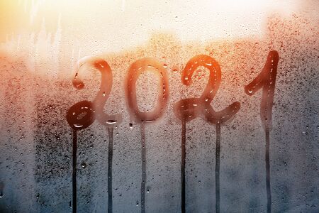 numbers 2021 of new year paint with finger with streaks of water on splashed by rain foggy glass on blue window and sunsetの写真素材