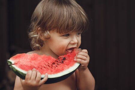 baby boy 2 years holds and bites piece of ripe red watermelon with seeds, selective focus on melon, copy spaceの写真素材
