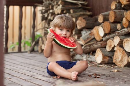 baby boy 2 years in blue shorts holds and bites piece of ripe red watermelon with seeds, selective focus on melon, copy spaceの写真素材