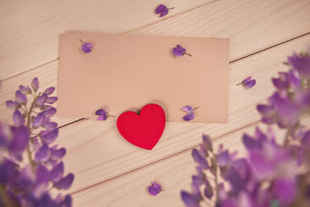 red wooden heart lies on brown envelope on rustic table in frame of flowers lupine and scattered...