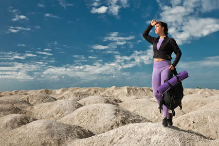 sporty young woman solo hiker on hilly desert in sportswear and with backpack holding sports looking into the distanceの写真素材