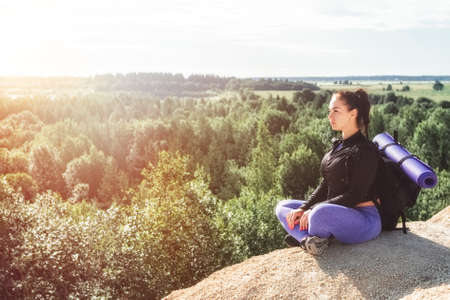 sports woman single hiker with backpack sitts on mountain in yoga pose meditation and looks into...