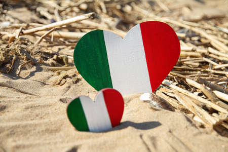 twain wooden heart painted in the colors of flag of Italy green, white and red lies on golden sand on sunny day beach, tourism concept, national day, selective focusの写真素材