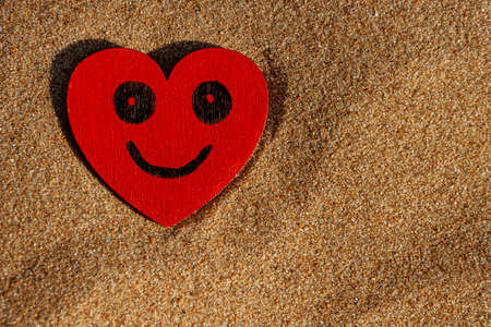 painted black smiling face on red wooden heart and yellow sand on beach on sunny day, love symbolの写真素材