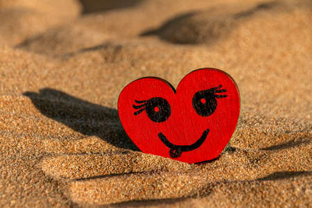 painted black smiling face on red wooden heart and yellow sand on beach, sunny day, love symbolの写真素材