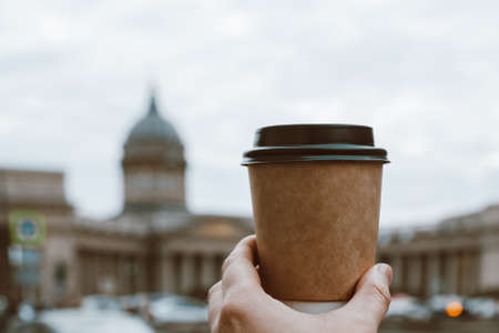 paper cup of natural coffee in womans palm on background of river Kazan Cathedral in St. Petersburg and sky, copy spaceの写真素材