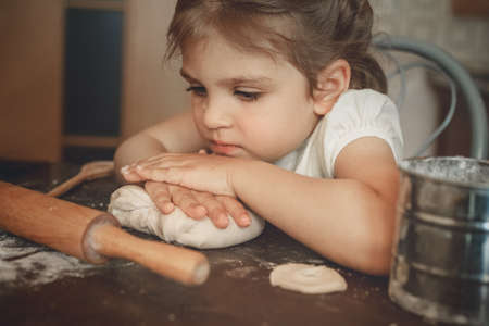 small child girl 3 years old sits at the table in kitchen thoughtfully put her hands ondough, next to there isrolling pinの写真素材