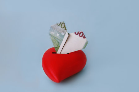 100 euro banknote rolled into heart shape inserted into red heart moneybox on blue background, copy spaceの写真素材