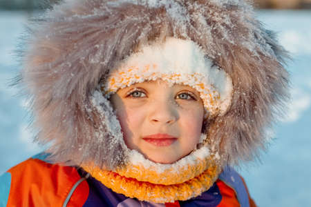 child girl 3 years old in winter jacket, yellow hat and scarf covered with snow, close-upの写真素材