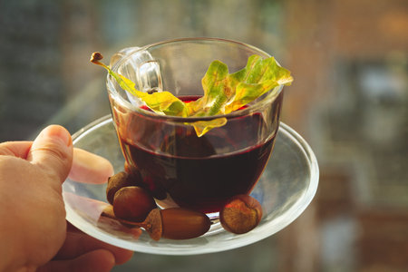 glass cup with tea with oak autumn leaf and acorns in womans hand, selective focusの写真素材