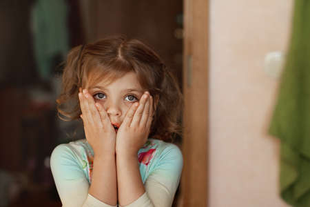 Close-up portrait of surprised little girl child with fcovered her face with her palms at home,...