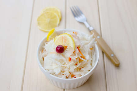 sauerkraut with carrot and cranberries, lemon, metal fork in white ceramic cup on light wooden table, selective focusの写真素材