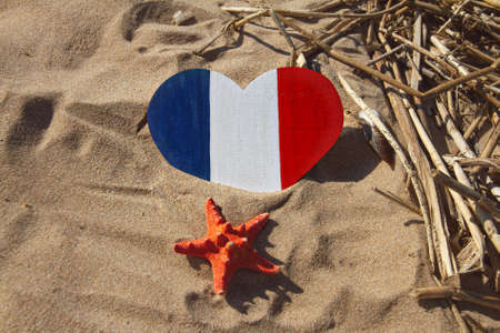 wooden heart painted in colors of the flag of France blue, white and red stands on golden sand with straw and shells on beach on sunny day, tourism concept, national dayの写真素材