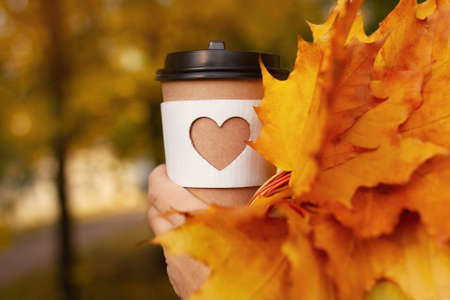 cup of coffee with carved white heart and yellow autumn maple leaves in womans palm on yellow autumn...