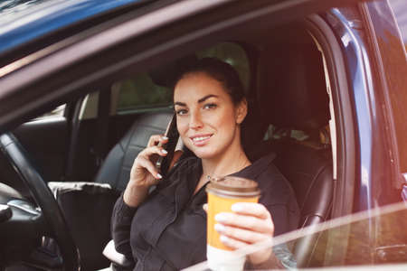 smiling brunette woman sitting in car with cup of coffee and smartphoneの写真素材