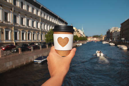 paper cup of natural coffee in womans palm on background of Moika river in St. Petersburg in summer...