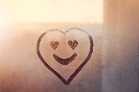 Happy smile heart shaped facepainted on window flooded with raindrops on blur red glass background in sunset cityの写真素材
