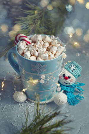 winter mug with marshmellow and knitted handmade stand snowman in blue scarf against background of festive light bokeh and fir branches, selective focusの写真素材