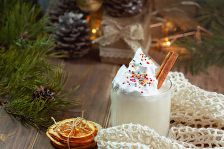 glass of white eggnog with cream and colored decoration on wooden brown table with spruce branches, mesh bag and dried orange slices, selective focusの写真素材