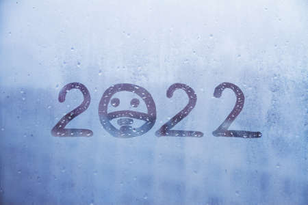 Handwritten blue arabic new Year numerals 2022 with sad face in protective medical mask on misted glass on window flooded with raindrops backgroundの写真素材
