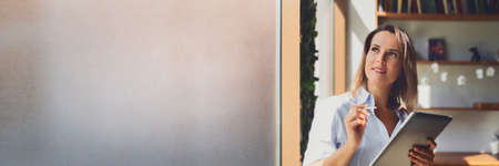 Portrait of smiling young woman in blue shirt with e-book tablet next to window in blur, banner,...