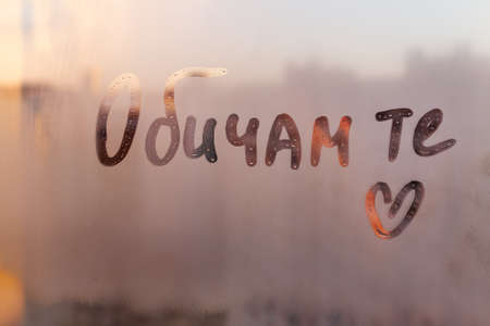 Handwritten message in Bulgarian language words I love you in english and hand drawn heart on misted glass on sunset window flooded with raindropsの写真素材