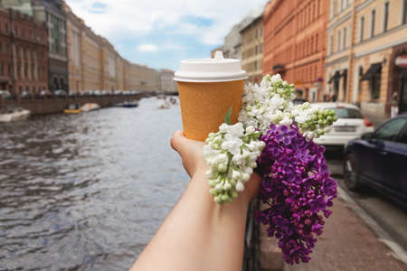 brown natural paper cup of ground coffee in woman's hand with flower of lilac on background of river European city on sunny dayの写真素材