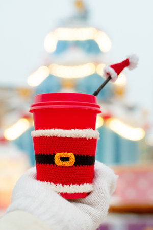 one red beige cups with white lid dressed in santa claus clothes in palm of hand in white mitten against background of festive multi-colored Christmas lightsの写真素材