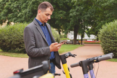 Portrait of middle-aged white man in business suit in phone rents kicksharing scooter on city street