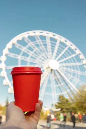 red natural paper cup of ground coffee in woman's hand on background of ferris wheel in European cityの写真素材