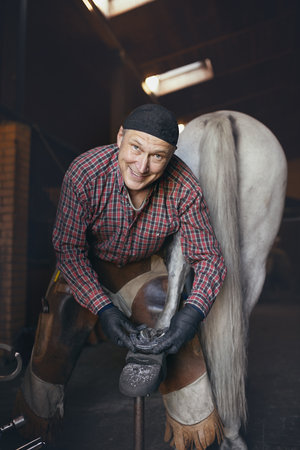 portrait of happy smiling European blacksmith shoves back hoof of white horse, selectve focusの写真素材