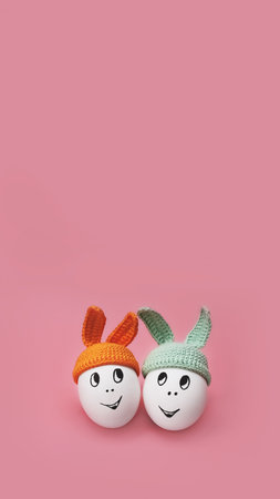 banner two white eggs in green and orange beanie with rabbit ears and cheerful faces on pink background, copy space, vertical, 16:9の写真素材
