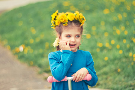 portrait of little dreamy girl in blue dress in wreath of dandelions with scooter, selective focusの写真素材