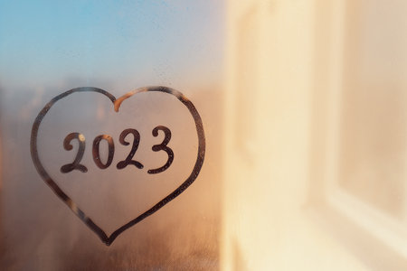numbers 2023 of new year in heart shape paint with finger on splashed by rain foggy sunset glass on windowの写真素材