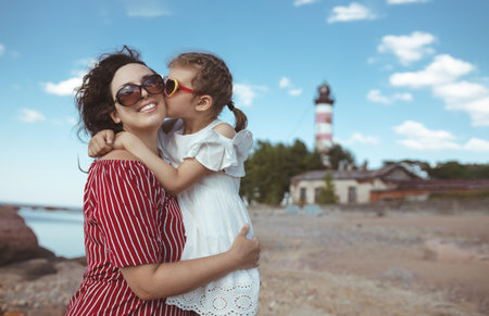 little girl of 5 years kisses happy mother in white and red dresses on beachの写真素材