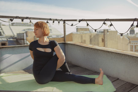Young woman in black sportswear sits green mat doing yoga and fitness exercises on terrace roof