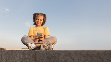 Girl 6-7 years old sitting on background of sky and playing on phone