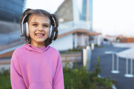Portrait of smiling baby girl 6-7 years old in pink hoodie listening with headphones and sits outdoor in cityの写真素材