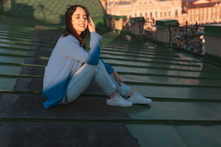 Portrait of young happy Caucasian woman in jeans and blue jacket sits resting on rooftop in St Petersburgの写真素材