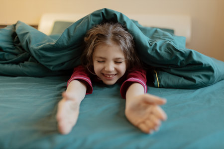 Happy 7 year old girl in red pyjamas in green blanket arms outstretched on bed at homeの写真素材