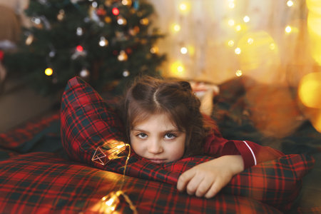 Child girl in red pajamas lies on festive bed with garland next to Christmas tree waiting for holiday at homeの写真素材