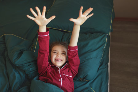 Happy 7 year old girl in red pyjamas in green blanket arms outstretched on bed at homeの写真素材