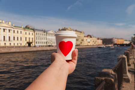 white paper cup of natural coffee with red heart in womans palm near embankment of Fontanka river in...