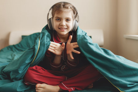 Happy 7 year old girl in red pyjamas and headphones sits in green bed with blanket at homeの写真素材