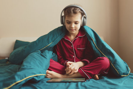 7 year old girl in red pyjamas and headphones sits in green bed with blanket and looking at tablet at homeの写真素材