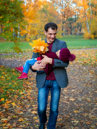 Happy daddy carries his baby daughter in his arms in autumn park with bouquet of yellow leavesの写真素材