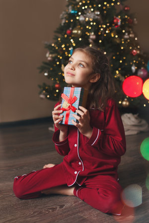 Happy girl of 7 years old in red pajamas sits on living room floor with gifts box next to Christmas treeの写真素材